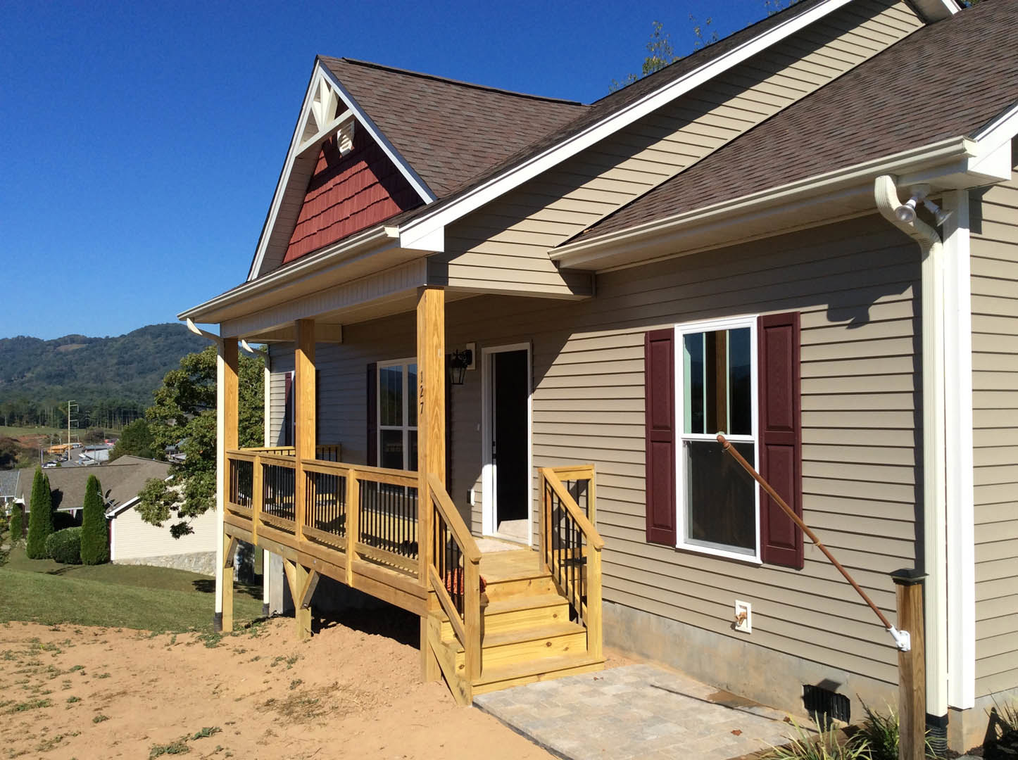 White cottage-style home with wooden porch and stairs, black front door with white frame, large windows, wooden deck railing, surrounded by tall trees and landscaped yard.