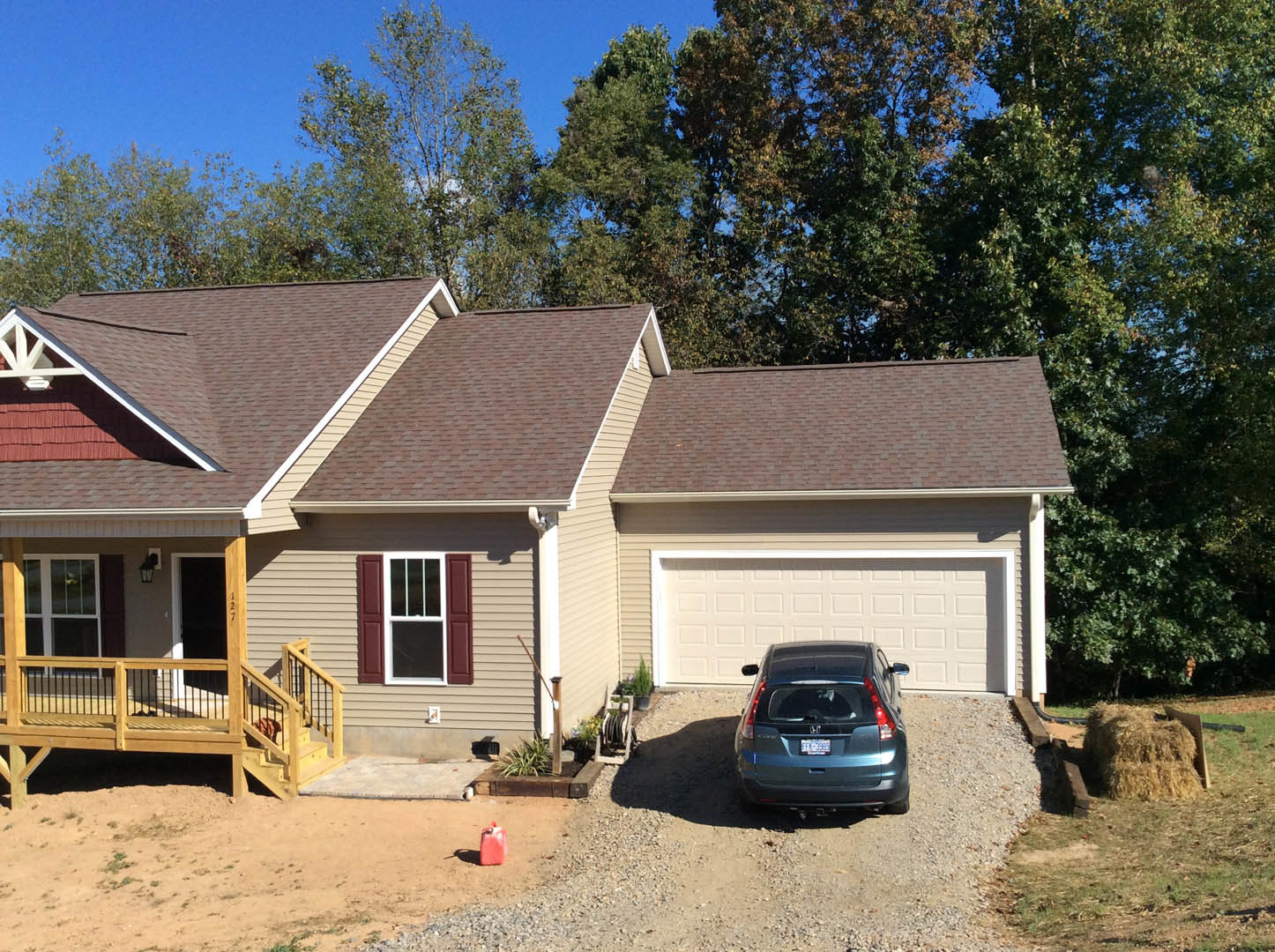 Two-story house with light siding, attached garage, wooden porch with railing, parked car in driveway, mature trees and blue sky in background