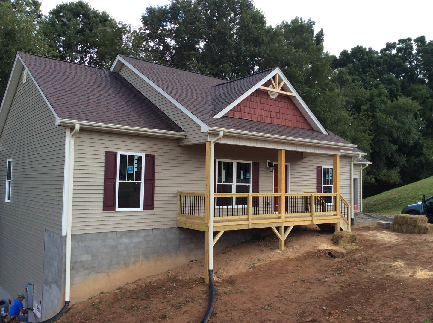 White siding house with covered porch, wooden deck and railing, double front doors, garden hose coiled near entry, large windows including one with a sign, mature trees in