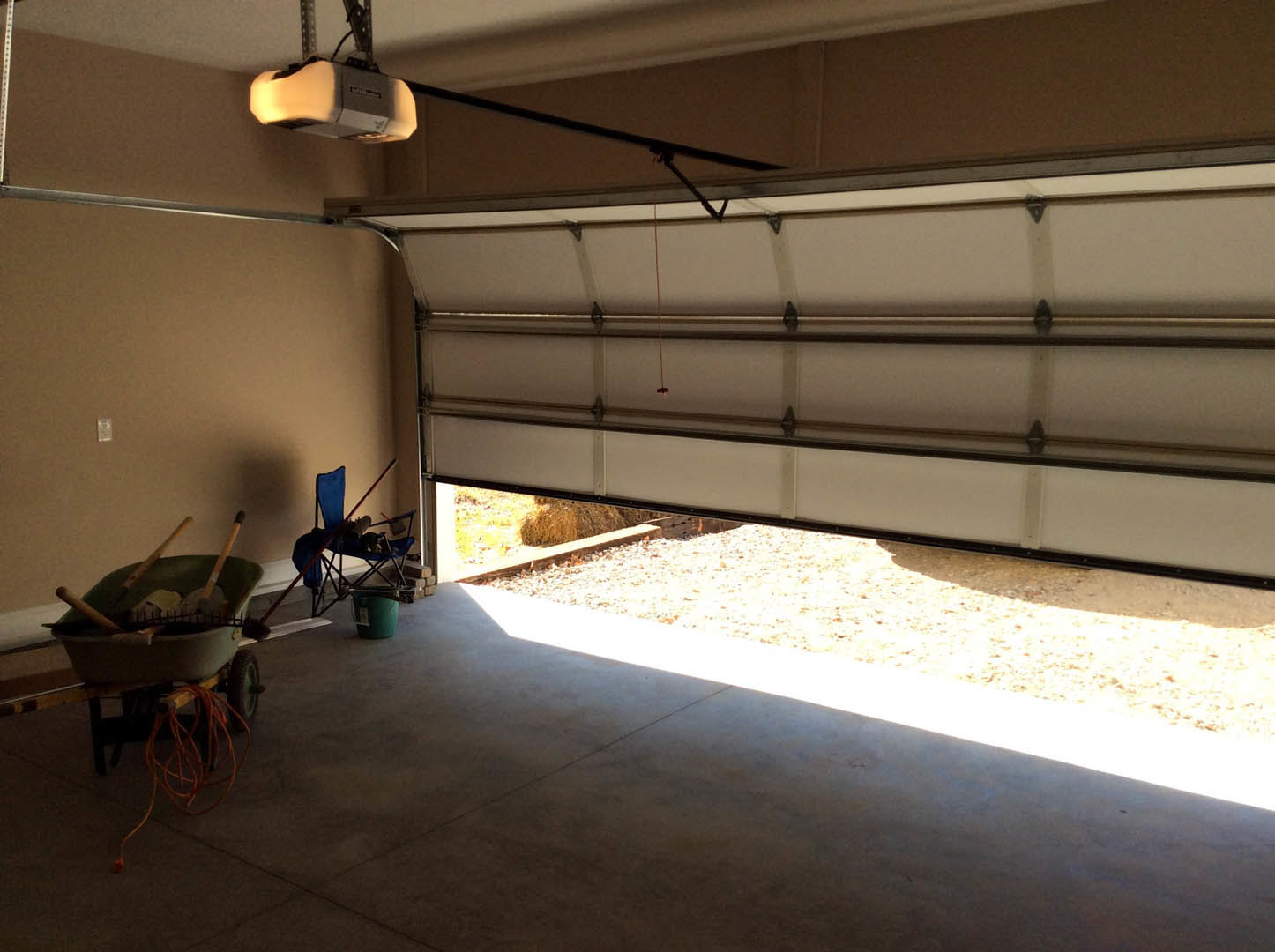 Garage interior with concrete floor, wheelbarrow filled with tools, wheelchair, blue folding chair draped with fabric, green bucket, and ceiling-mounted projector