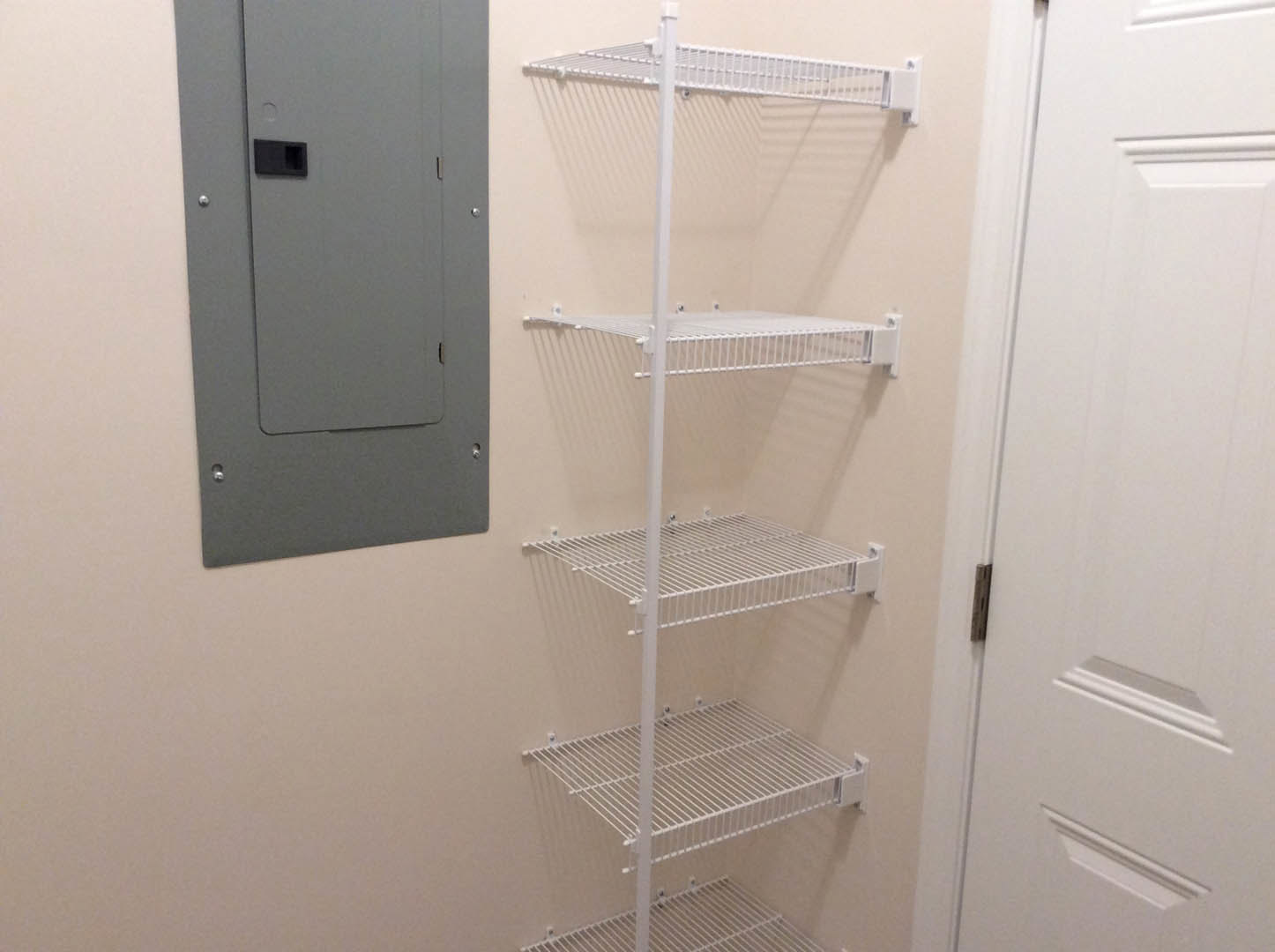 White built-in shelves on a wall beside a gray metal door with a square black handle, white corner shelf, and white bathroom shelf with metal grate and hinged white door.