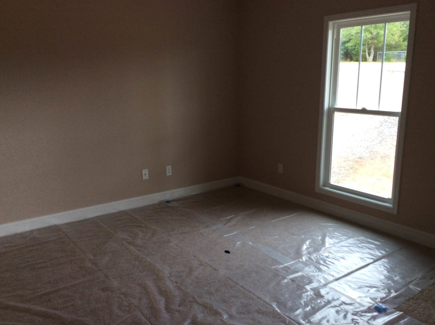 Sunlit room with white-framed window, plastic wrap covering laminate wood flooring, plastered walls, and leafy trees visible outside