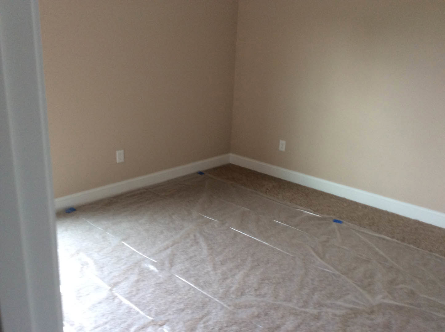 Carpeted floor covered with clear plastic sheeting, white walls, blue painter’s tape securing the plastic along the baseboard