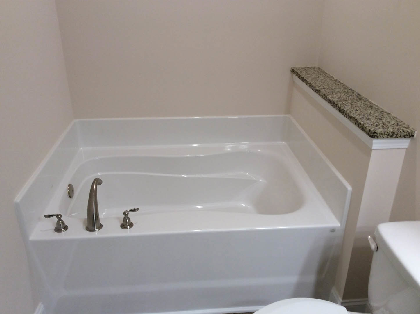 Freestanding white bathtub with chrome faucet and handle, granite shelf, and adjacent toilet on light tile floor