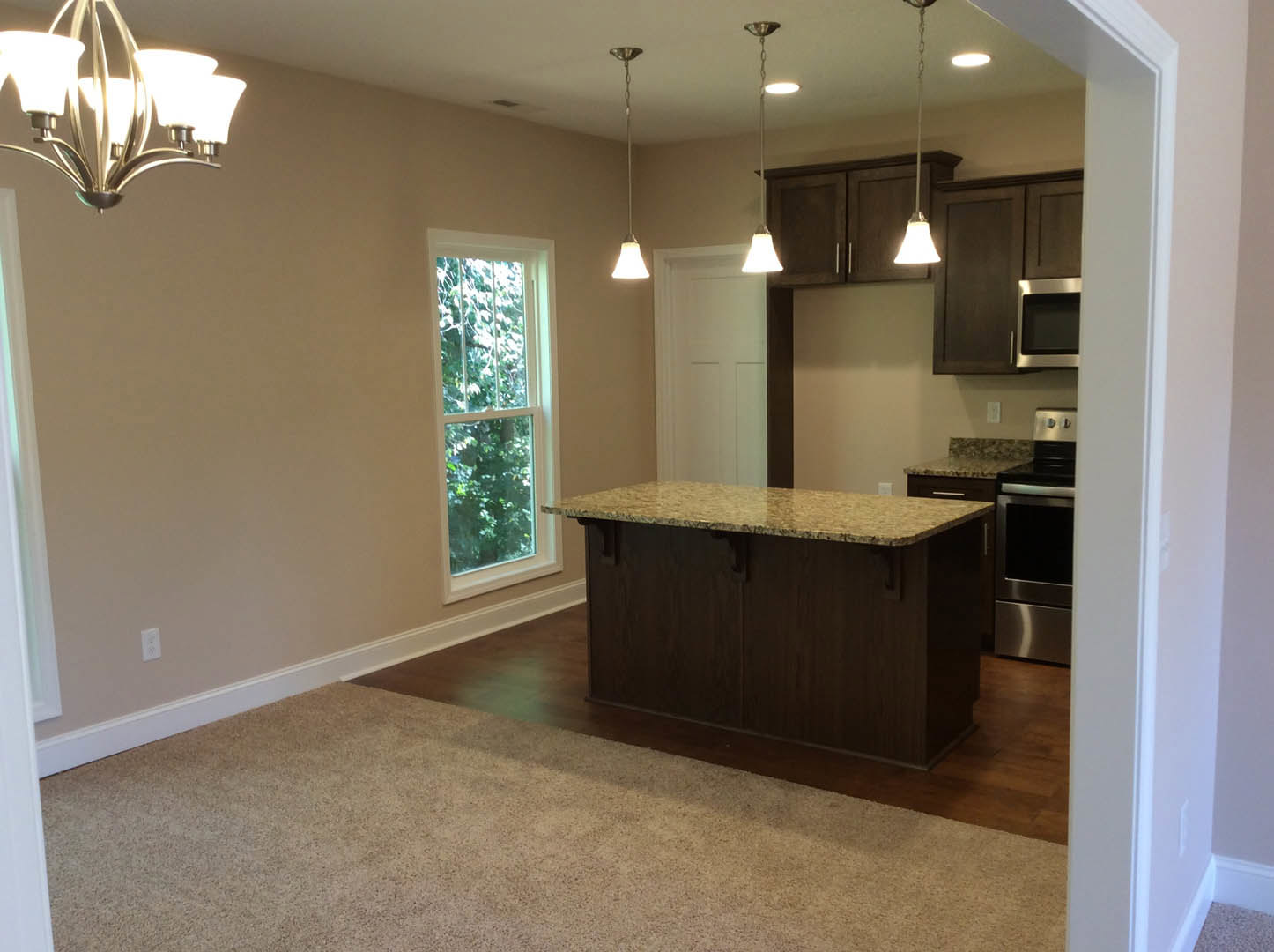 Open-concept kitchen featuring a bar with quartz countertop, stainless steel stove, white cabinetry, modern chandelier, built-in microwave, large window overlooking trees, and