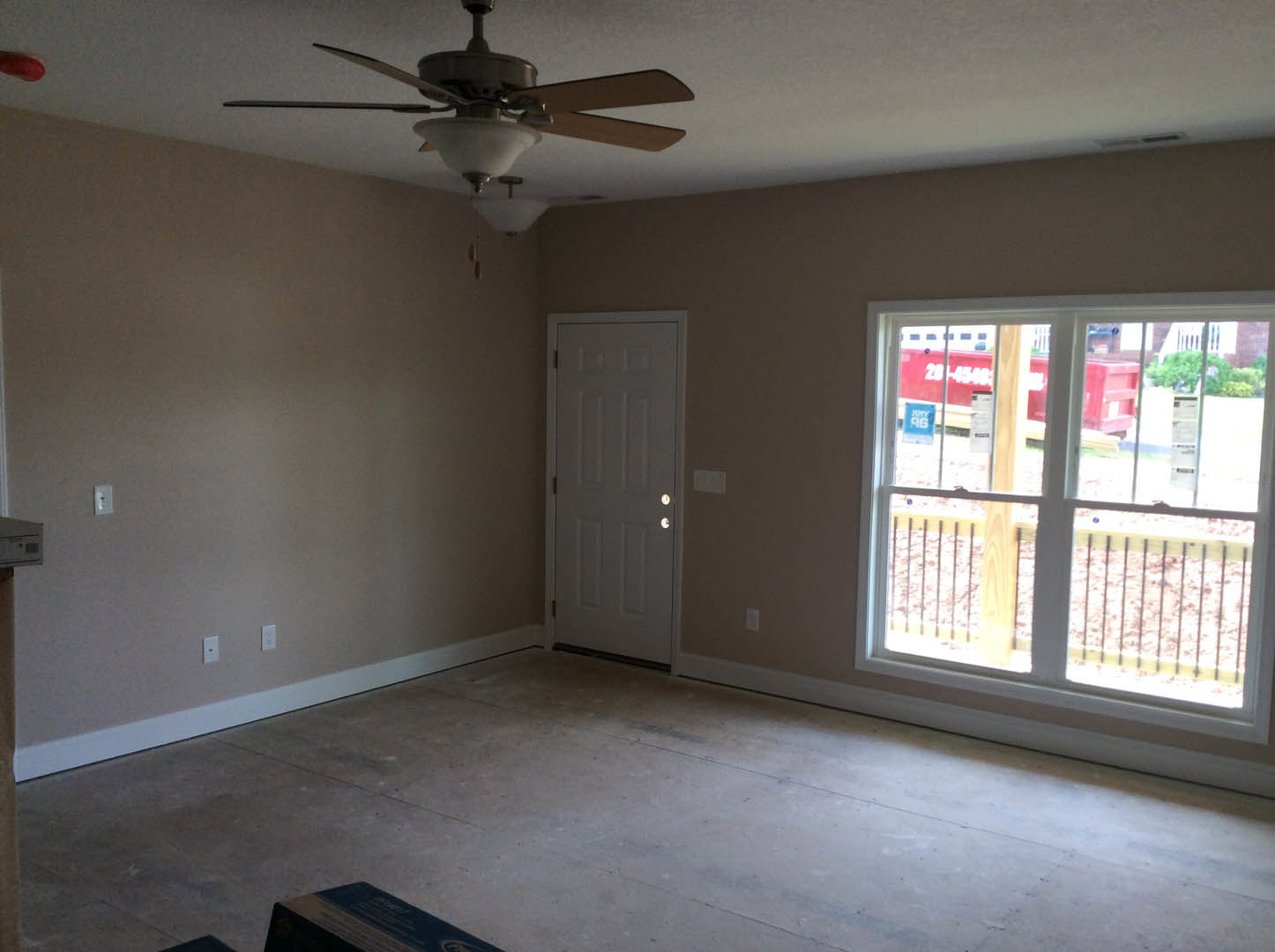 Neutral-toned room with plaster walls, wood flooring, ceiling fan with light fixture, single window showing exterior railing, white door, and a book placed on the floor