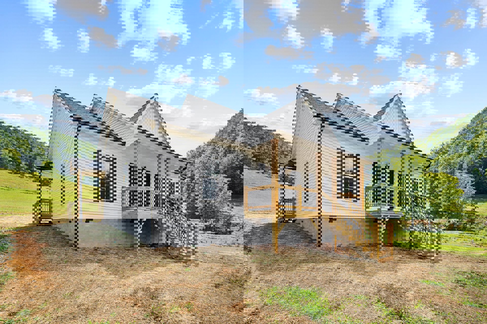 White cottage-style house with covered porch, wooden deck, exterior stairs, white-framed windows, grassy yard, and blue sky with scattered clouds