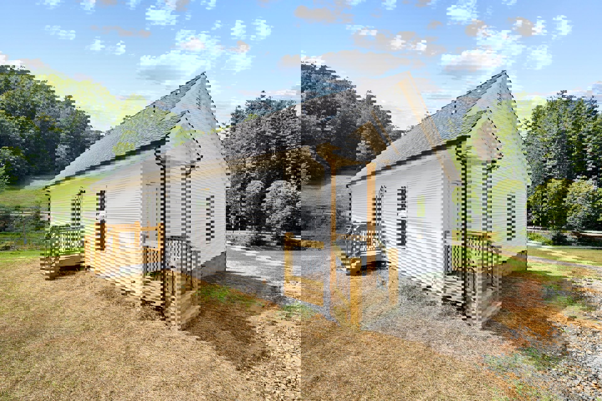 White siding house with wooden porch and black railing, stairs leading up to entry, surrounded by grass and trees under a partly cloudy blue sky.