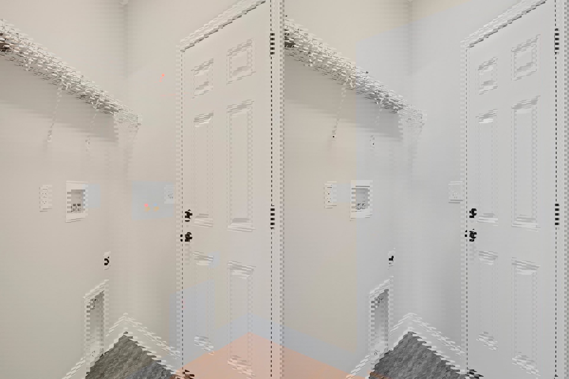 White paneled door with black knobs, white built-in shelf, wood flooring with white trim, electrical outlet, white-framed valves, and a roll of paper in a white box against plaster