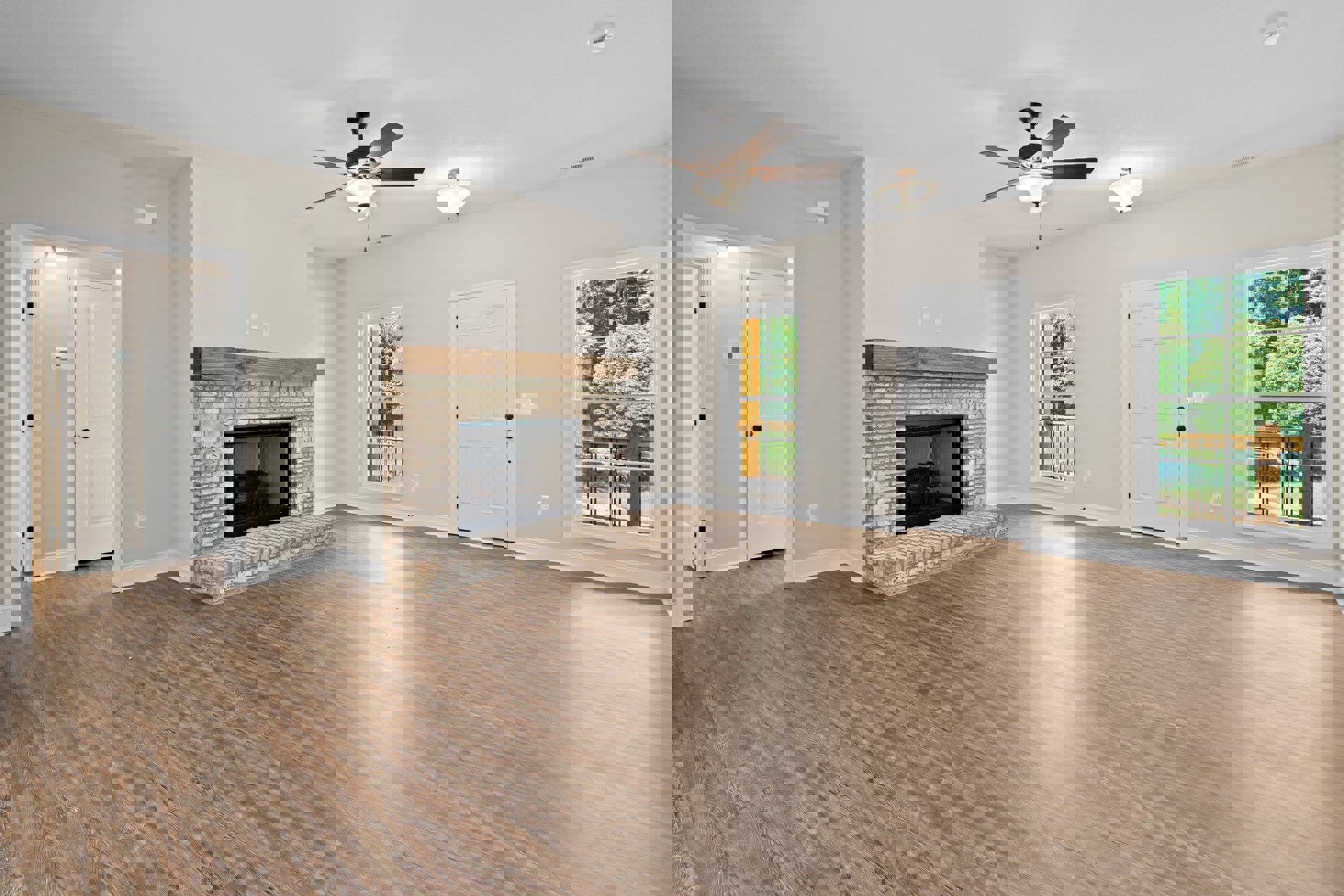 Living room with wood flooring, glass-door fireplace set in a white wall, ceiling fan with light fixture, white door with black knobs, window overlooking trees