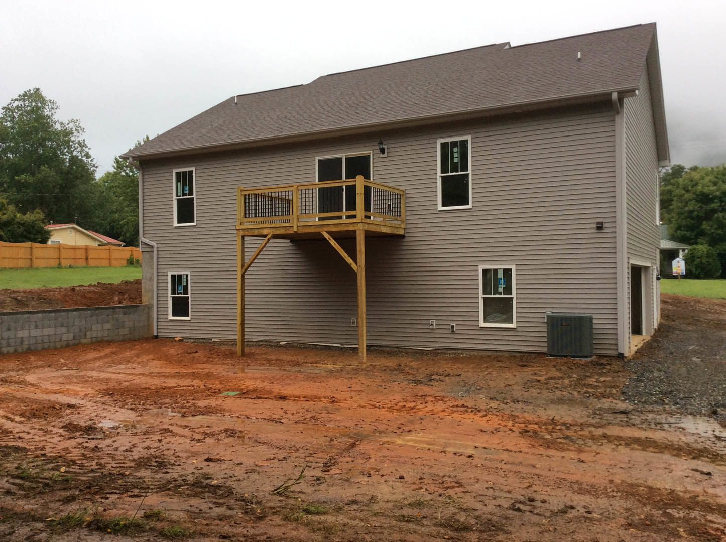 Two-story house with white-framed windows, wooden balcony and deck, dirt road and utility pole in front, trees and sky in background