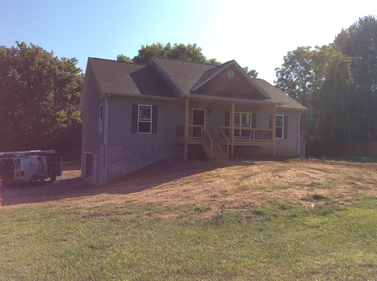 Two-story house with covered porch and stairs, white window frames, dirt hill with grass in foreground, trees in background, white van with ladder parked nearby