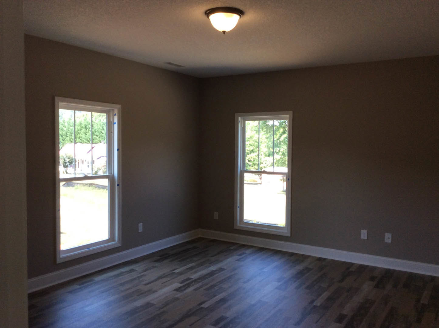 Sunlit room featuring large windows, hardwood flooring, white baseboards, recessed ceiling light, and views of trees, grass, and a neighboring house.