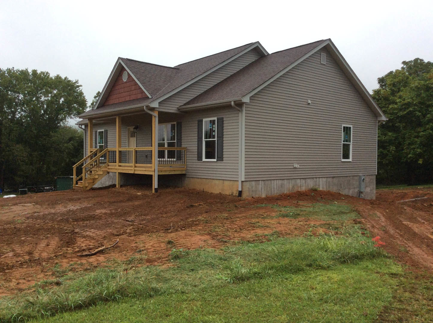 Two-story home with white-framed windows, wooden porch featuring metal railings, dirt yard patch in front, surrounded by trees and greenery.