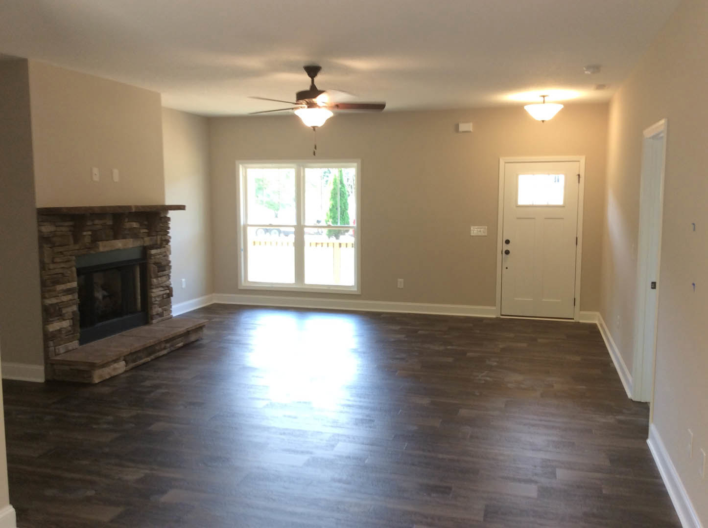 Living room with wood flooring, stone fireplace, ceiling fan, white-framed window, and white door with glass panel
