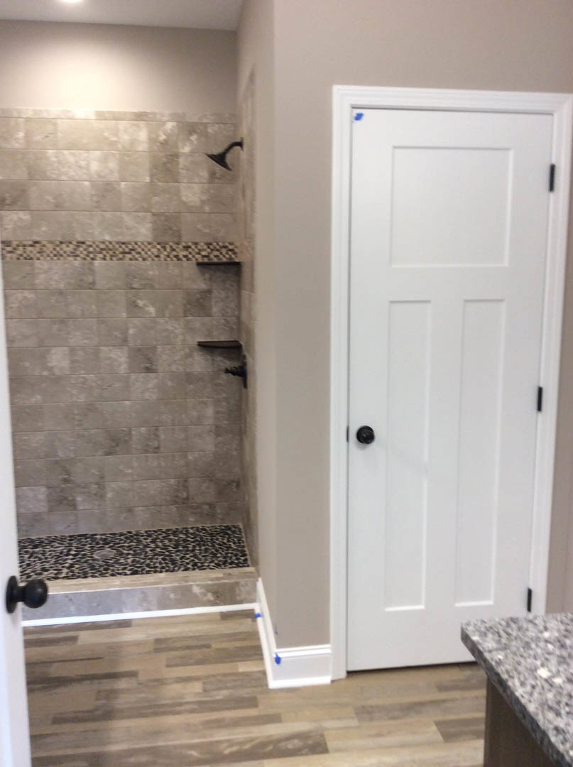 Bathroom with white door and black knob, wood flooring marked with blue tape, tile shower wall featuring a black shelf, and close-up of stone countertop; blurry bird visible in