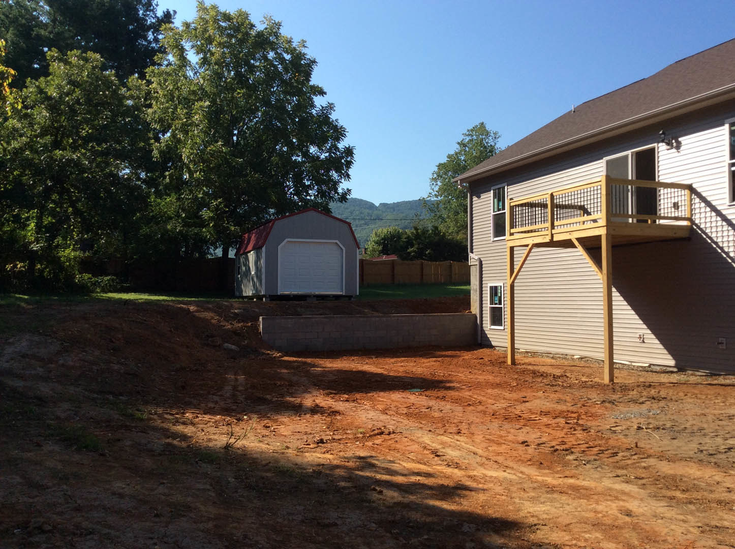 Two-car garage with white doors and frames, adjacent wooden deck, red-roofed shed, mature tree, dirt yard, and residential home exterior