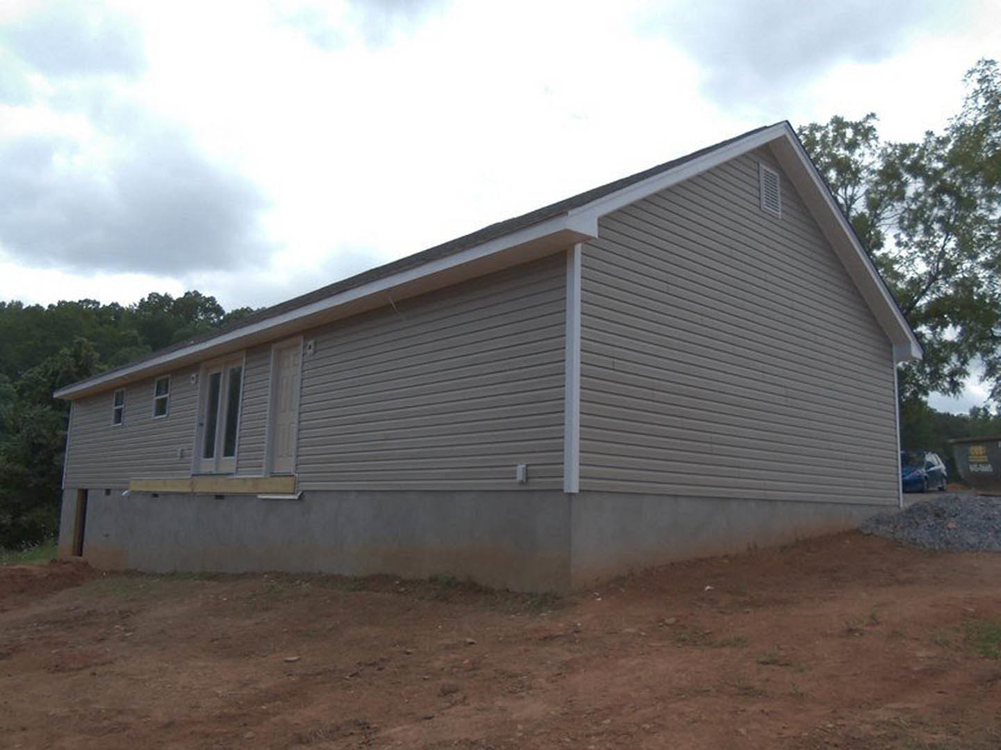 Partially built house with white door, exposed siding, dirt ground, pile of gravel, surrounding trees, and cloudy sky
