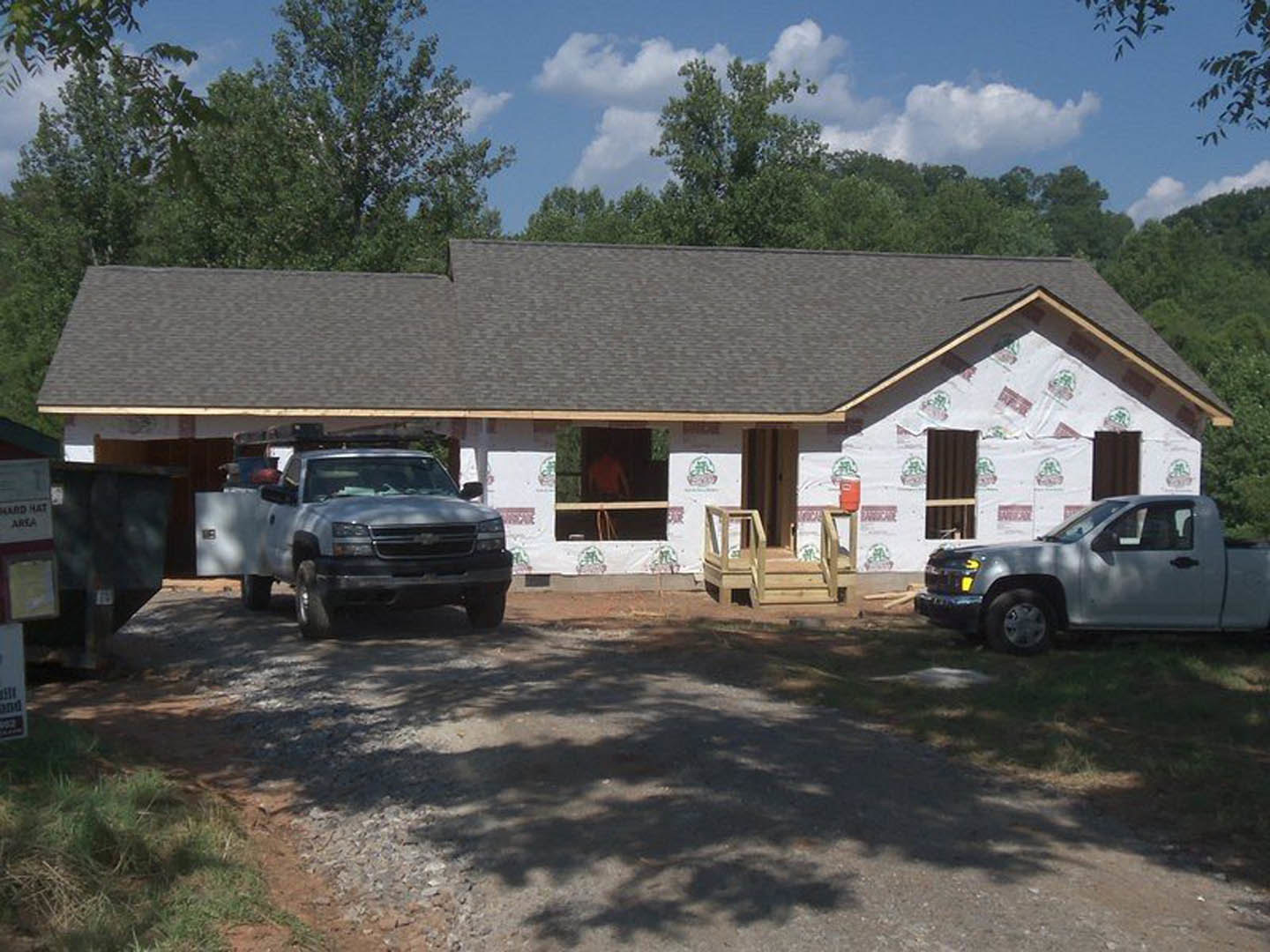 Two-story house framed with exposed wood and sheathing, construction truck with ladder parked in gravel driveway, cloudy sky overhead, trees in background.
