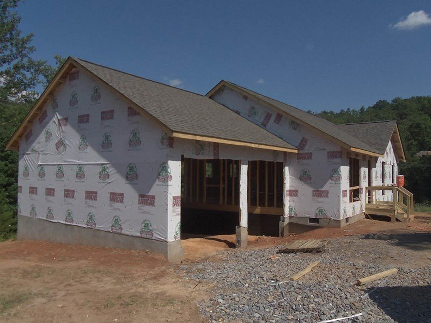 Framed house under construction with covered porch, exposed wooden beams, unfinished exterior walls, and roof installed
