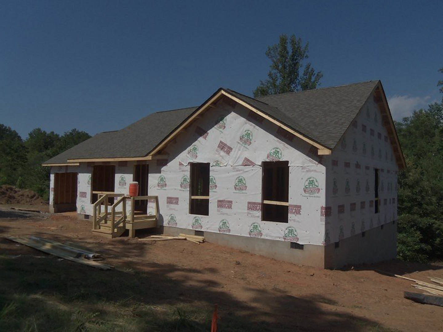 Partially built house wrapped in white plastic sheeting, exposed wooden framing, visible window openings, outdoor setting with trees and sky in background