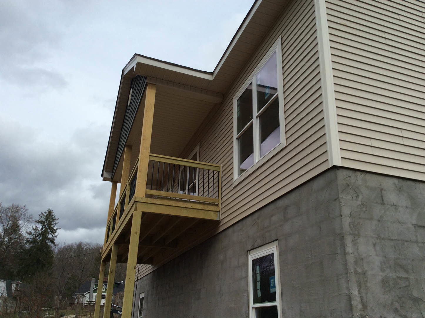 Two-story home with light siding, upper-level balcony with metal railing, large windows, and mature trees in the background