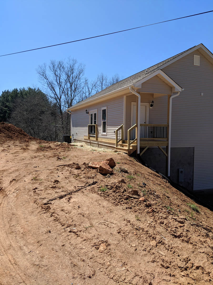 Two-story house with light siding, front porch and stairs, large dirt hill in foreground, leafless tree, power lines overhead, windows visible on facade