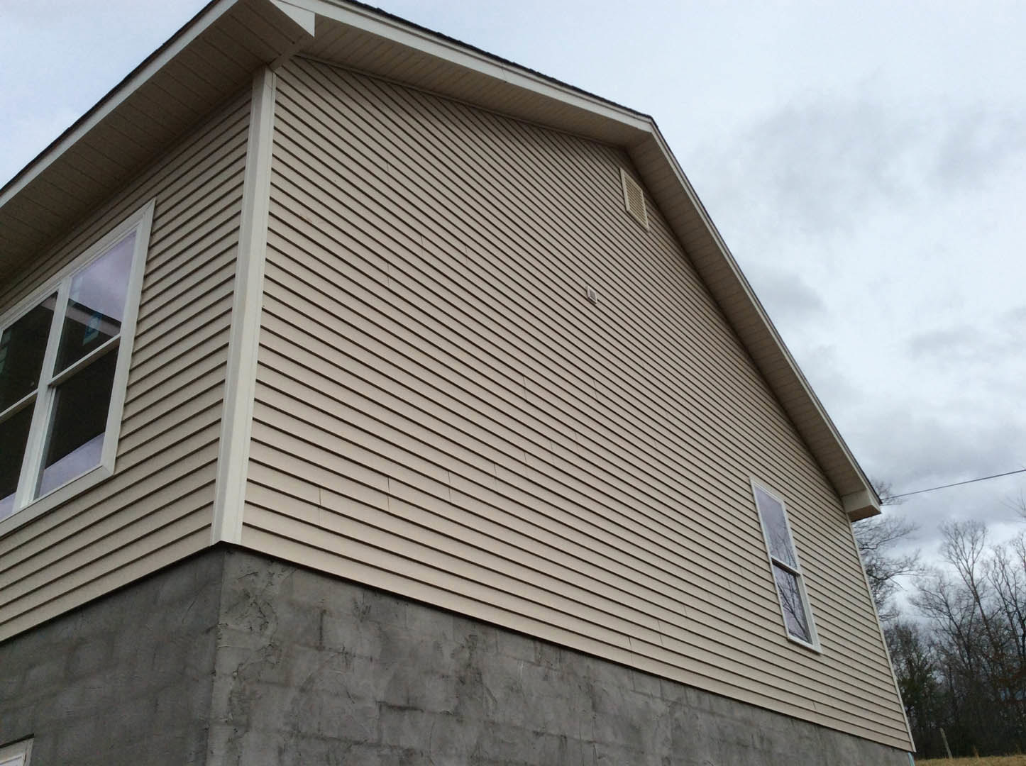 Modern home with grey siding, concrete foundation, rectangular window, roof vent, and tree branches against a cloudy sky