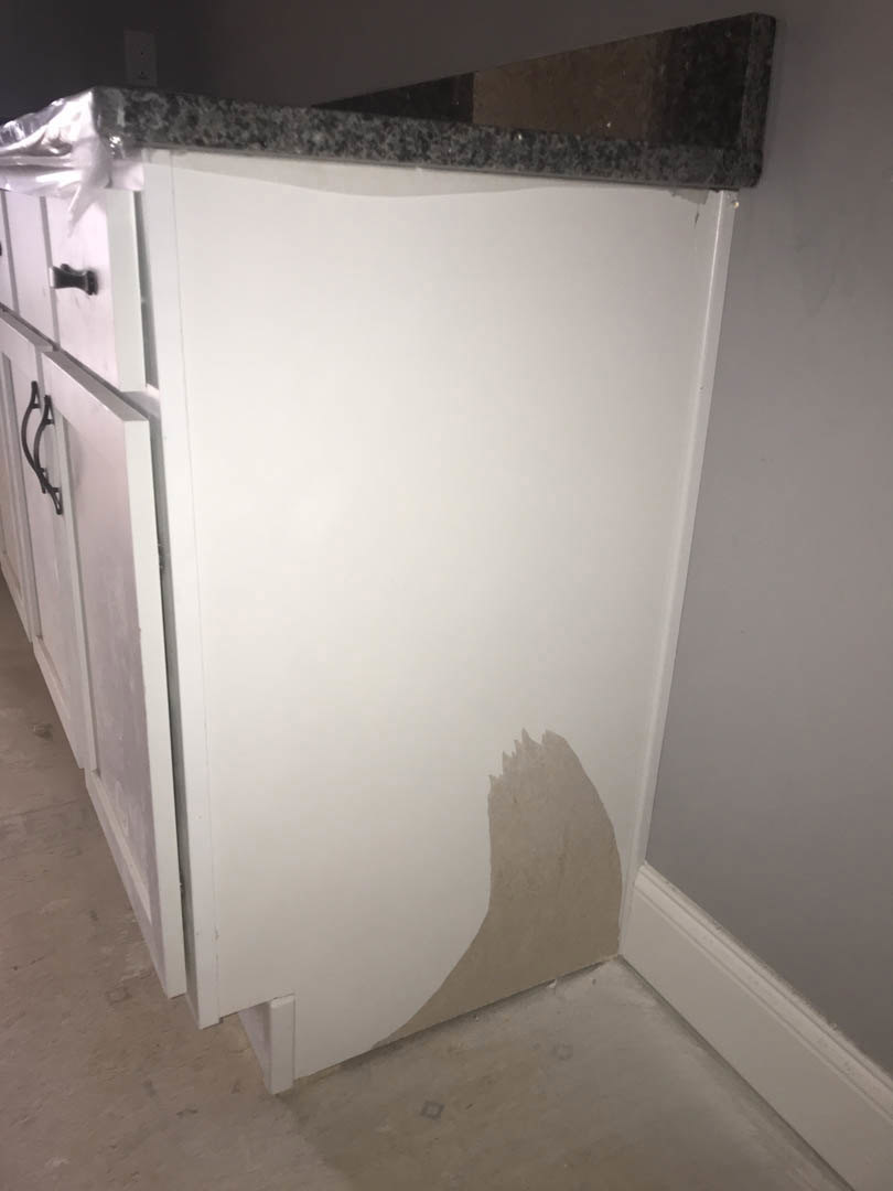 White kitchen cabinet with brown stain on corner, adjacent to plaster wall and light-colored floor, partial view of dishwasher appliance.