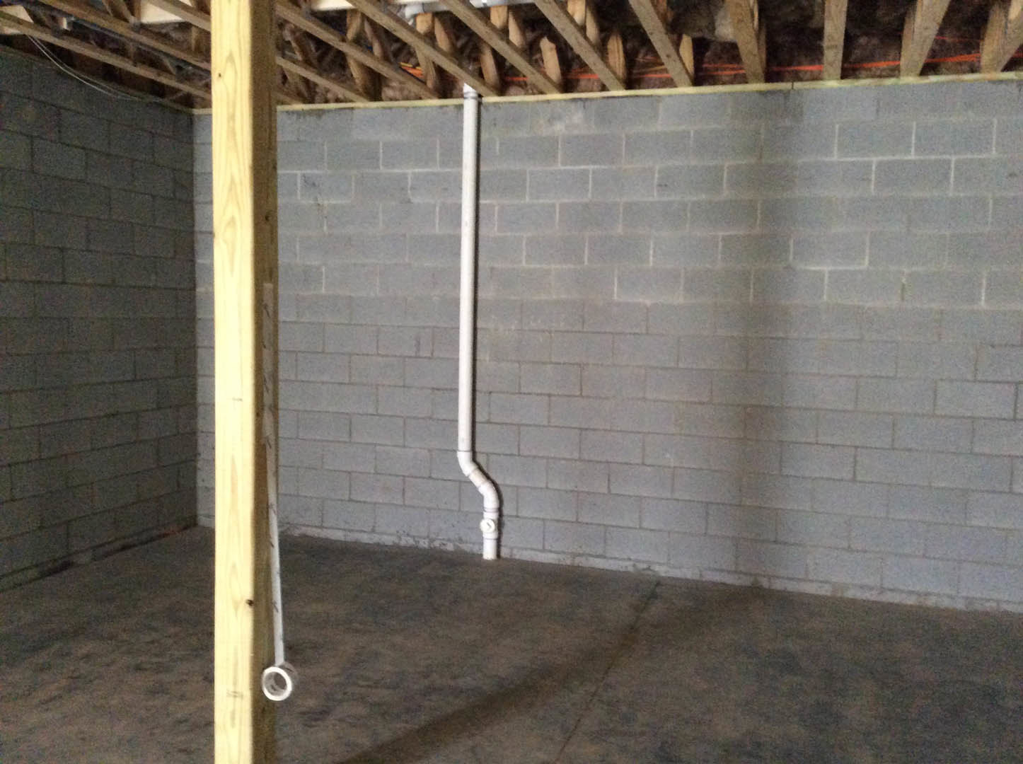 Exposed white pipe runs along a grey concrete wall beneath wooden ceiling beams in an unfinished room.