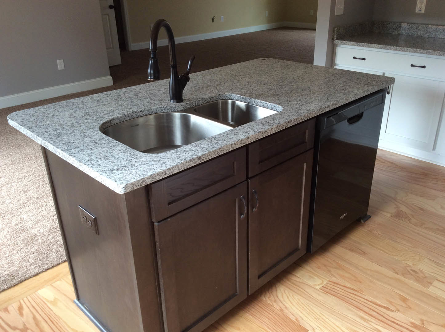 Granite kitchen island with stainless steel sink and modern faucet, brown cabinetry, black dishwasher, tile flooring, and wood surface reflections