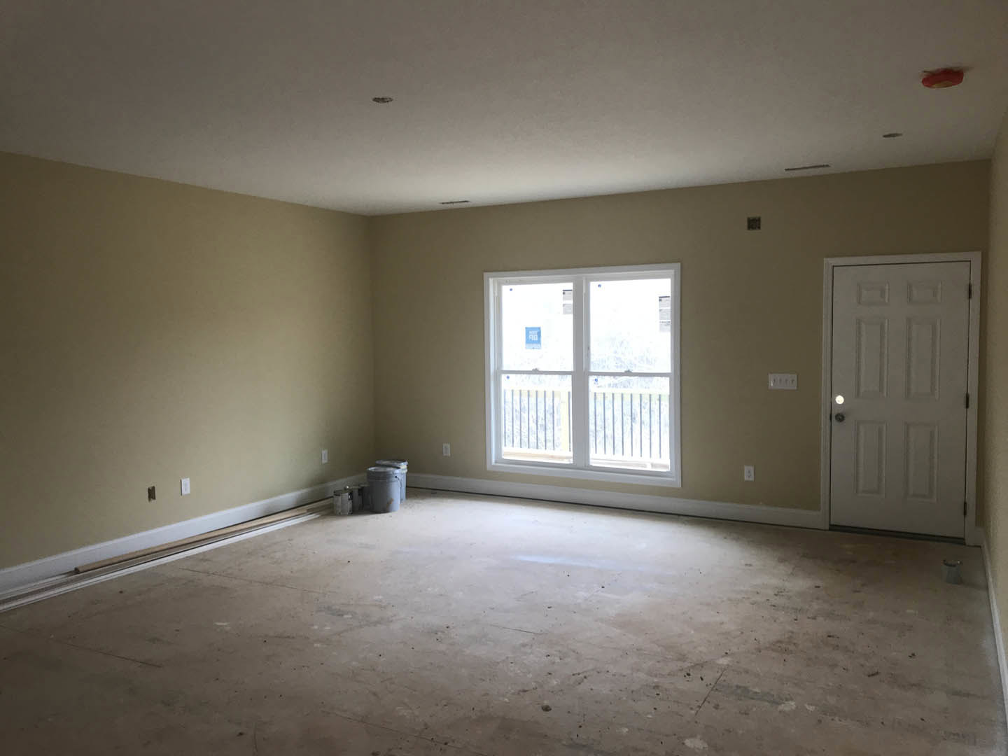 Unfinished room with white paneled door, window featuring blue sign, concrete floor with bucket, white plaster walls, and white ceiling with black and white fixture