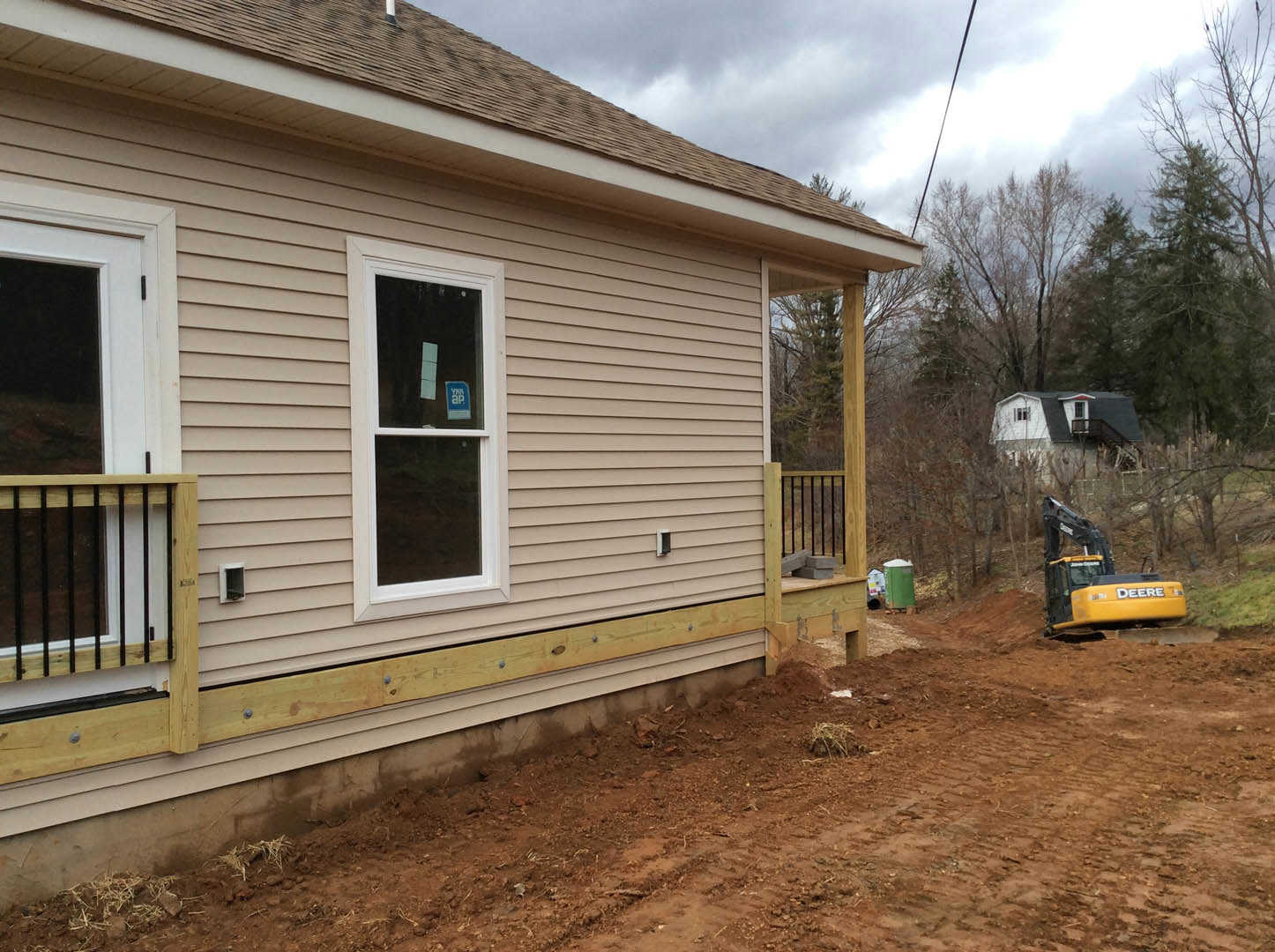 Partially built home with exposed framing, deck and railing, forklift parked in front, windows with signs and bars, siding installation in progress, small plant near foundation