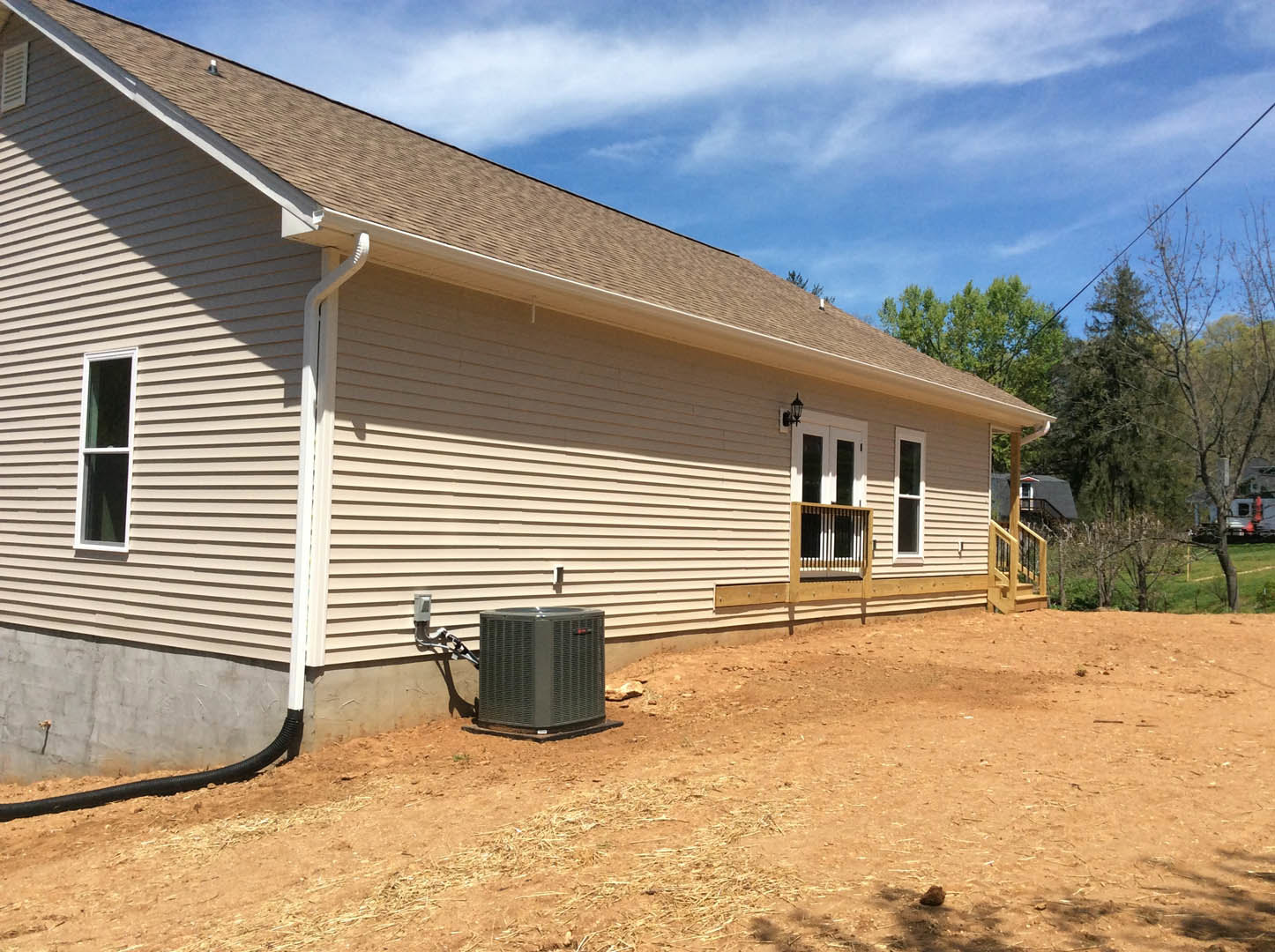 Grey heat pump installed beside white-framed window on house exterior with light siding, under roofline against blue sky with scattered clouds