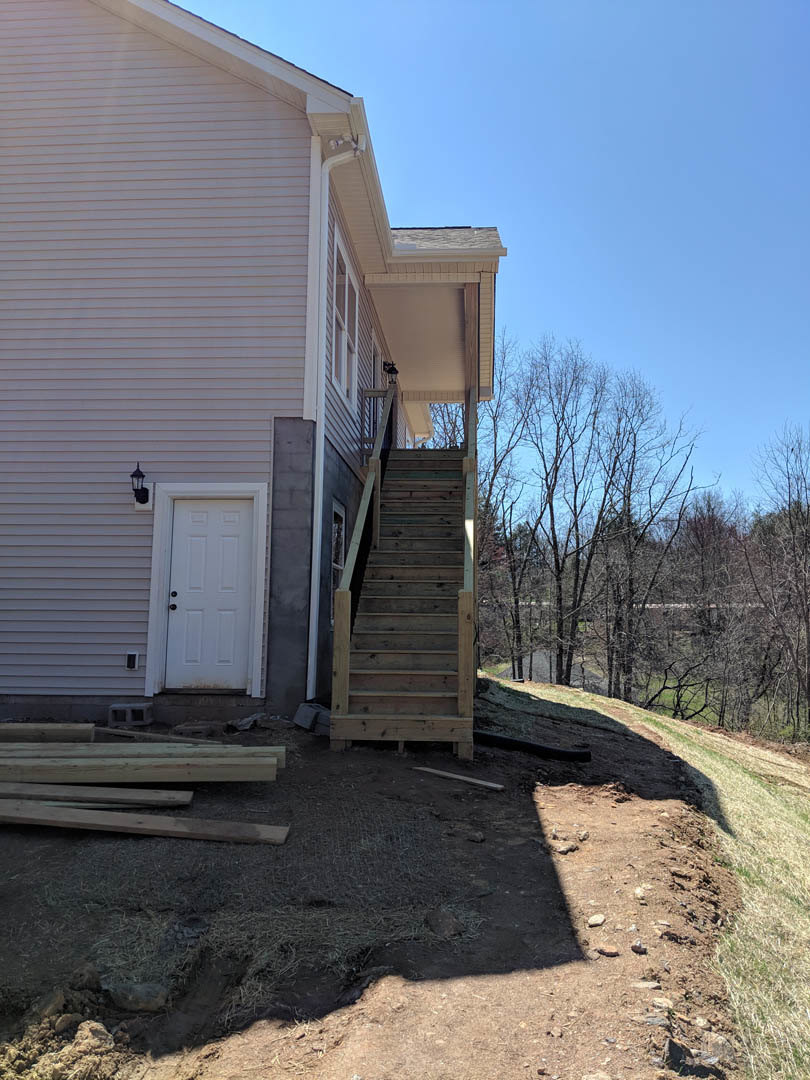 Wooden exterior staircase with natural wood planks leading to a white door with black hardware, surrounded by forest views and light-colored siding
