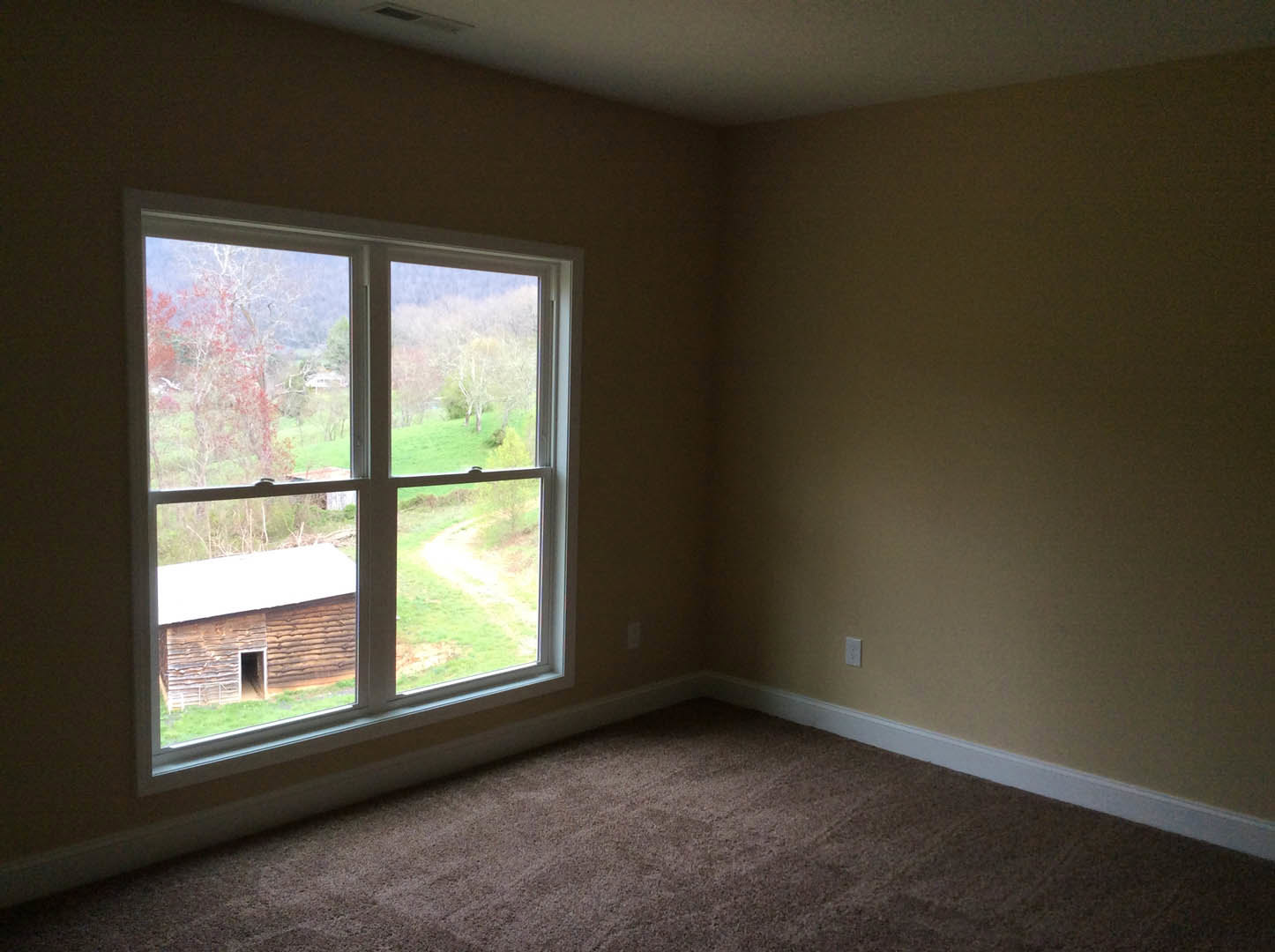 Carpeted room with a large window, white blinds partially open, grey painted walls, and a view of a log cabin and greenery outside.