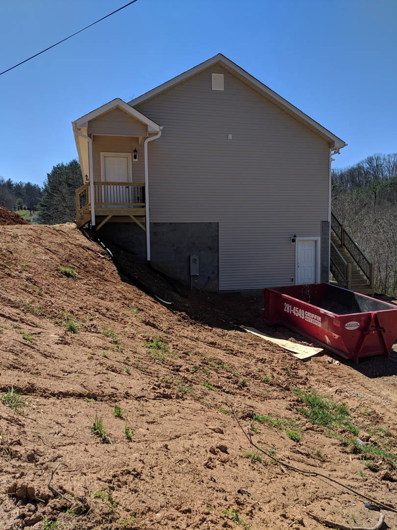 White house with side porch, red dumpster with white lettering on soil, grassy yard, white door with metal handle, trees and plants in background
