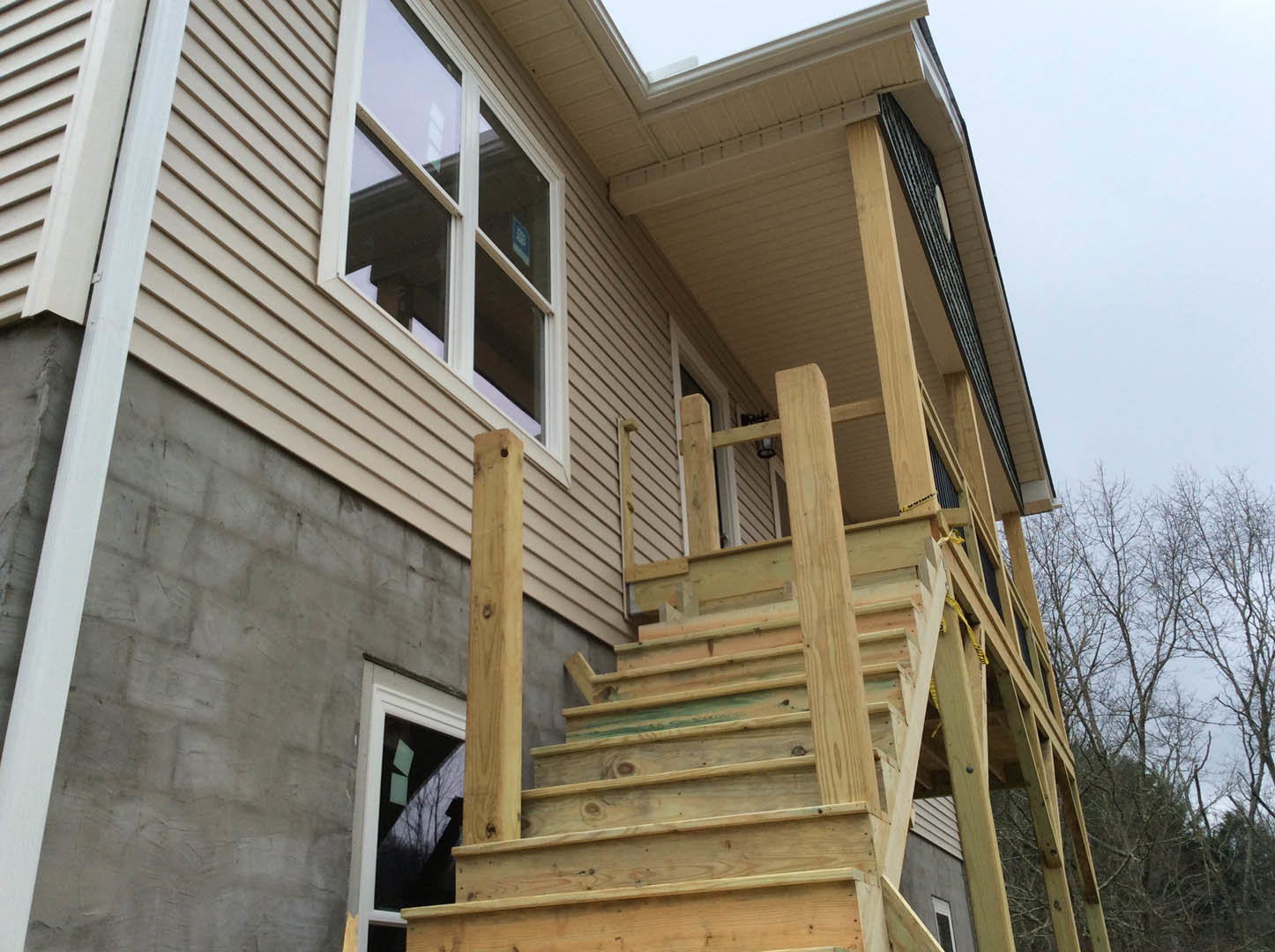 Wooden exterior stairs leading to porch, large window with tree branches reflected, light-colored siding, blue sky in background