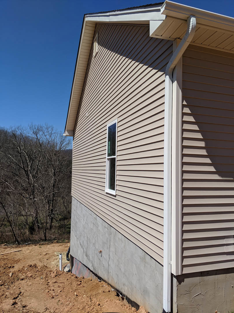 White siding exterior with rectangular window, exposed foundation, leafless tree nearby, dirt ground with visible pipe adjacent to wall