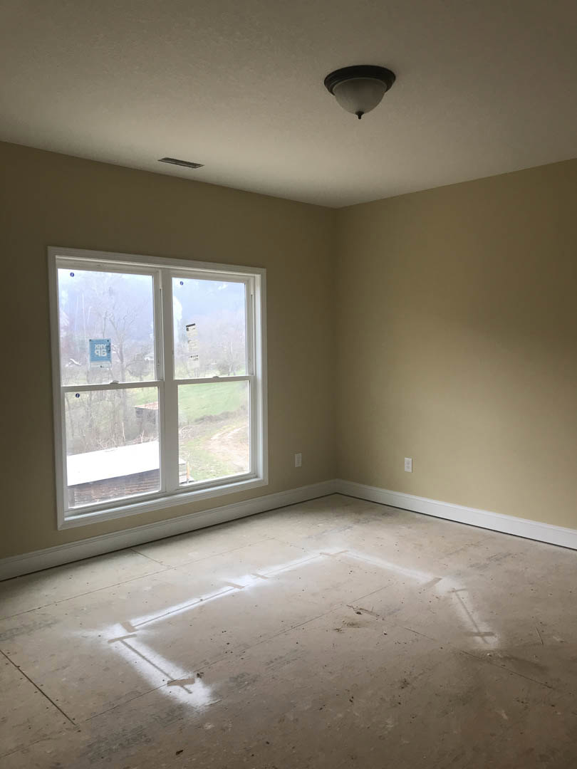 Sunlit room with white-framed window, plaster walls, ceiling light fixture, and tile flooring marked with white paint.