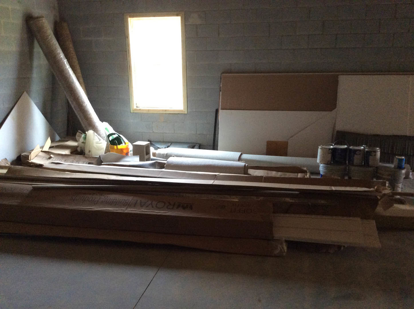 Cardboard boxes stacked on a wooden floor in a room with plaster walls, sunlight streaming through a window, wallpaper roll and jars visible in the background.