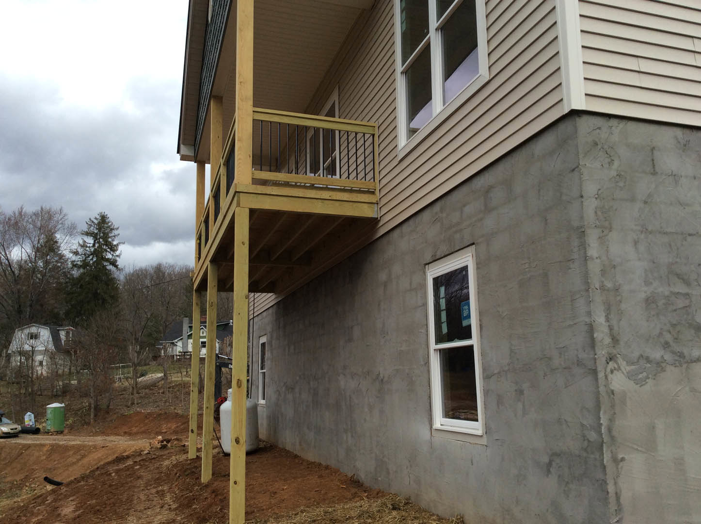 White two-story home with horizontal siding, second-floor balcony with metal railing, large windows with white frames, small porch area, leafy tree partially visible, blue sky and
