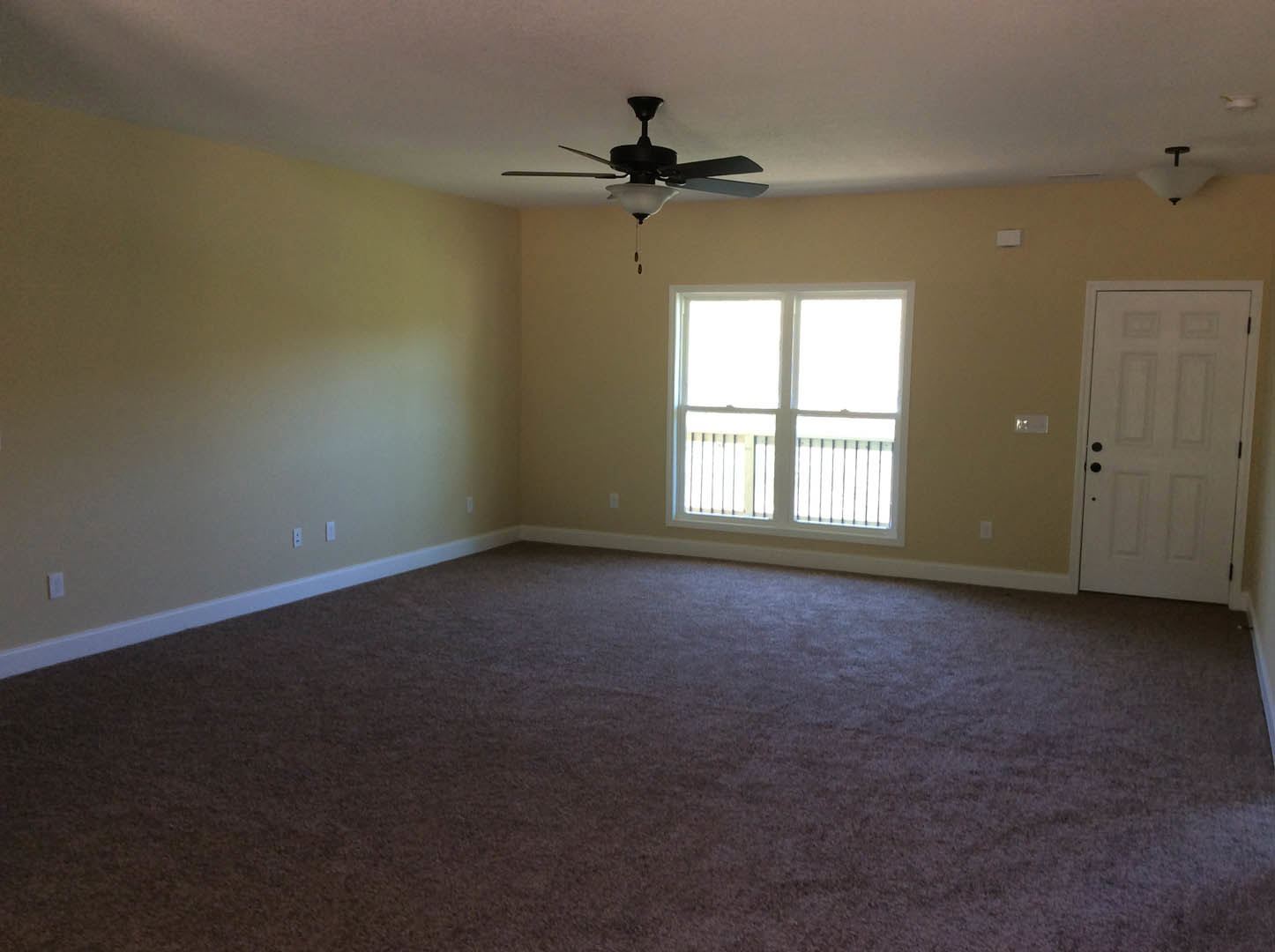 Bedroom with beige carpet, white walls, ceiling fan with light, white door featuring black hardware, and window with white railing