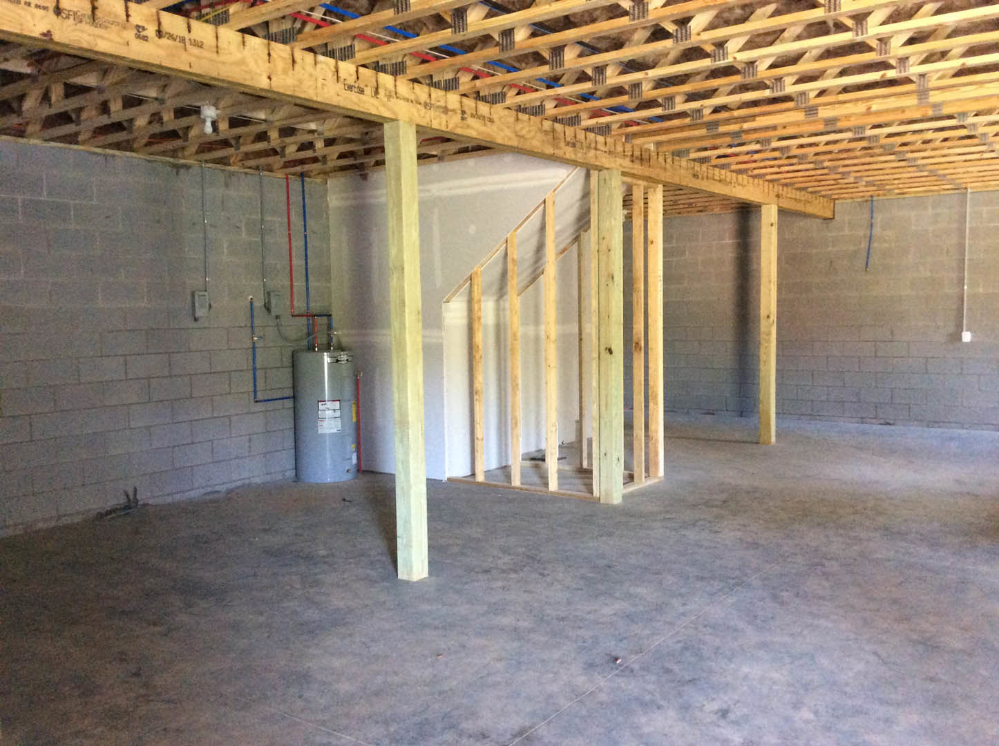 Basement room with exposed wood framing, concrete floor, large grey cylindrical tank with label, metal rods attached to wooden beam, and yellow object in foreground.