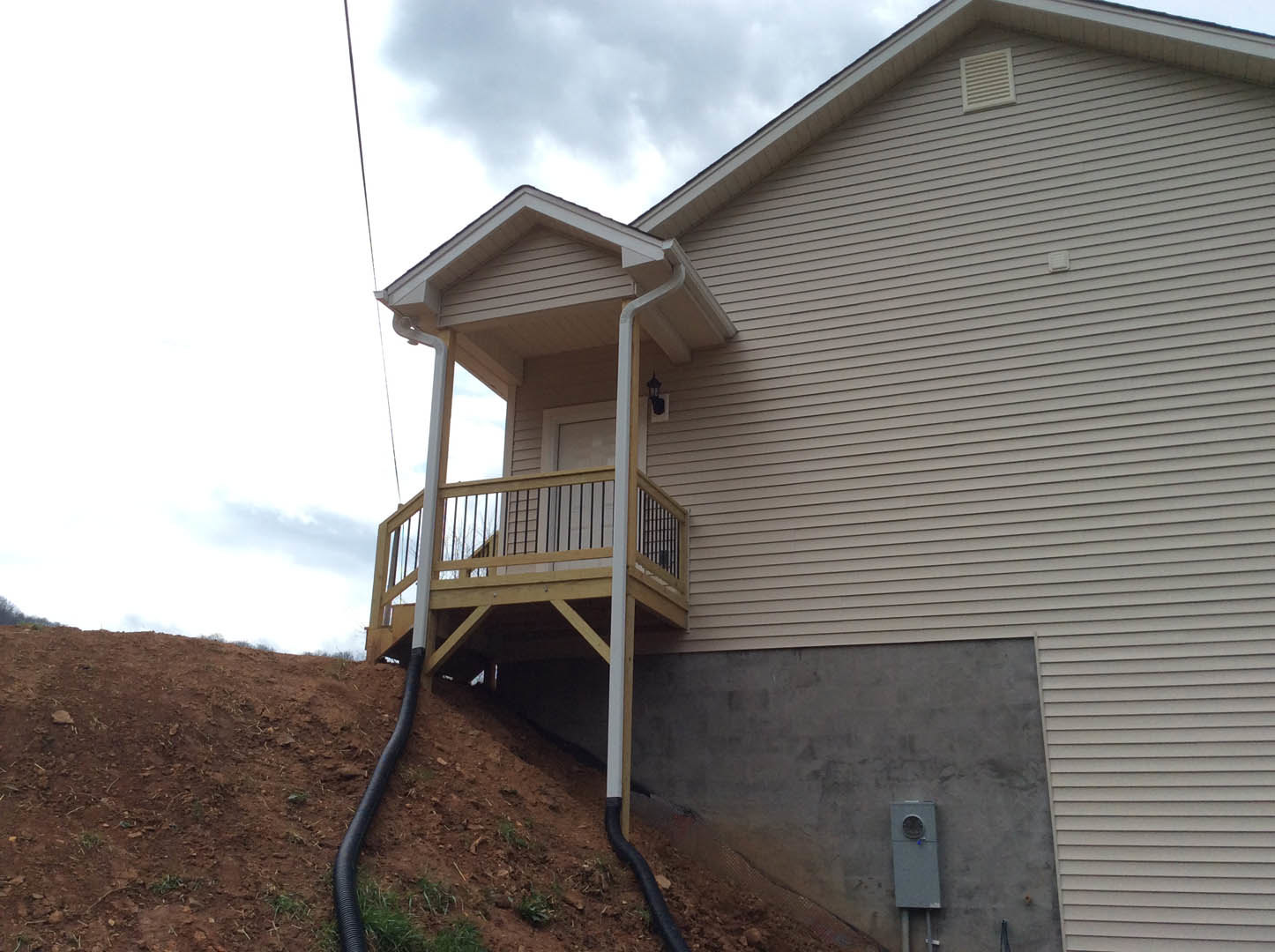 White siding house with covered porch, metal railing, and a vent pipe extending from the side wall; windows and roof eaves visible under partly cloudy sky.