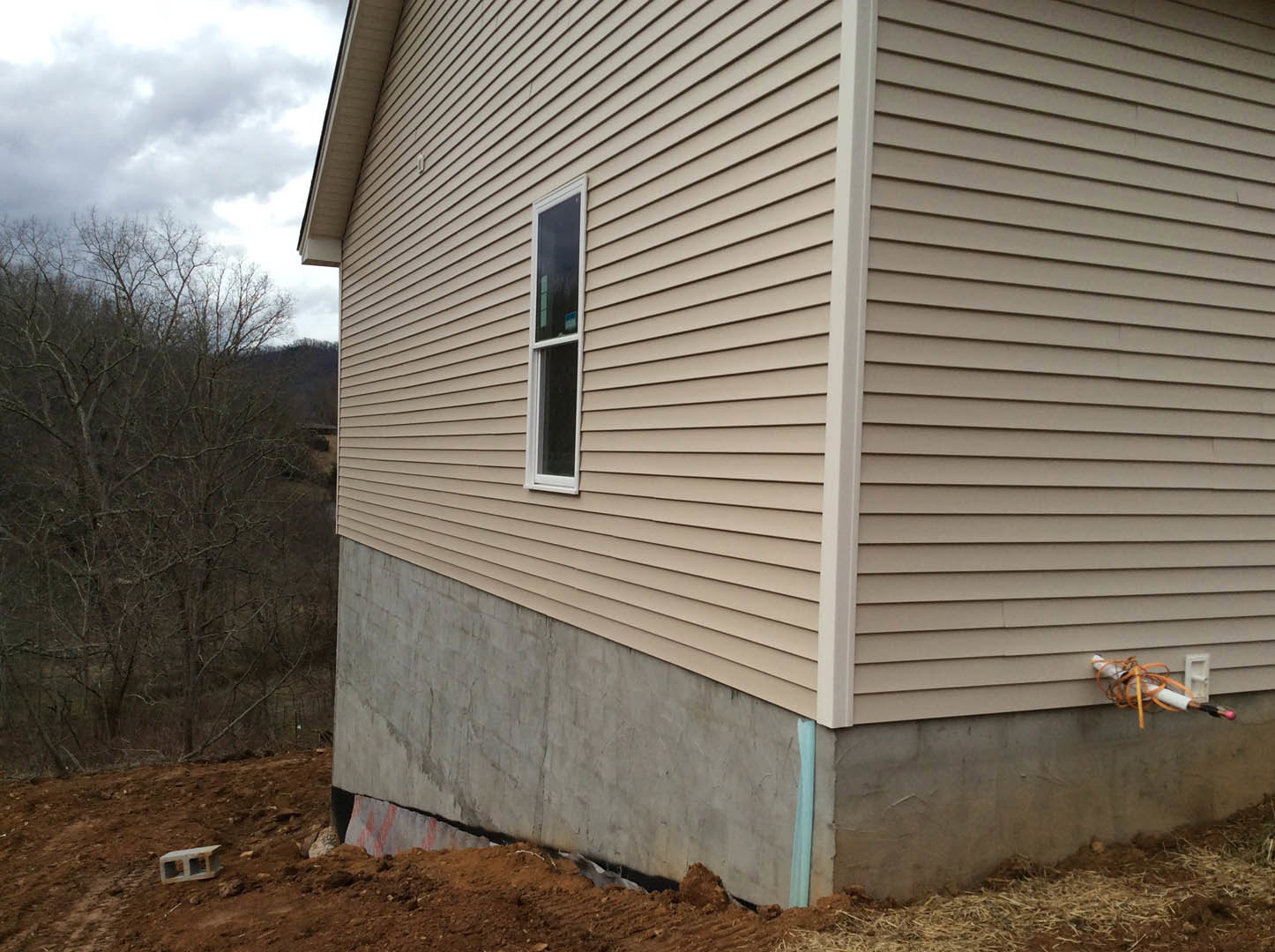 Partially built house with exposed concrete foundation, white-framed window, unfinished siding, leafless tree in background, cloudy sky overhead