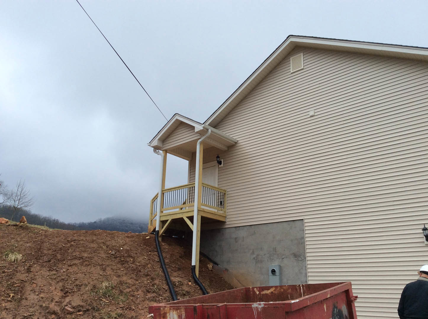 White house with covered front porch, construction dumpster on gravel driveway, cloudy sky overhead, man in blue jacket near porch, red paint container and vent visible, concrete