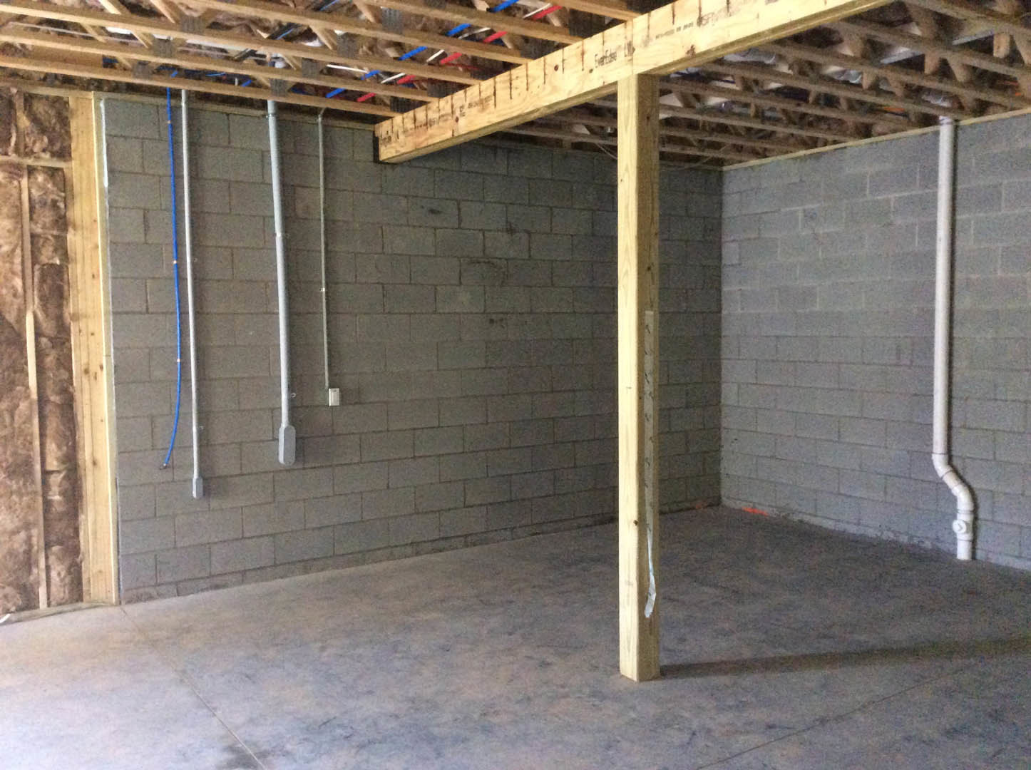 Open room featuring exposed wooden beam, red and blue pipes, concrete floor, and textured brick wall