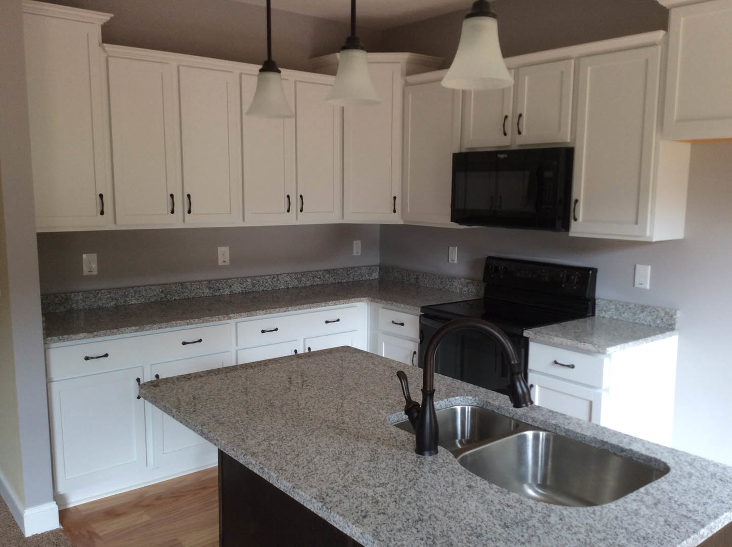 White kitchen cabinets with granite countertops, stainless steel sink and faucet, black microwave oven, ceiling light fixture, and lamp shade visible in foreground.