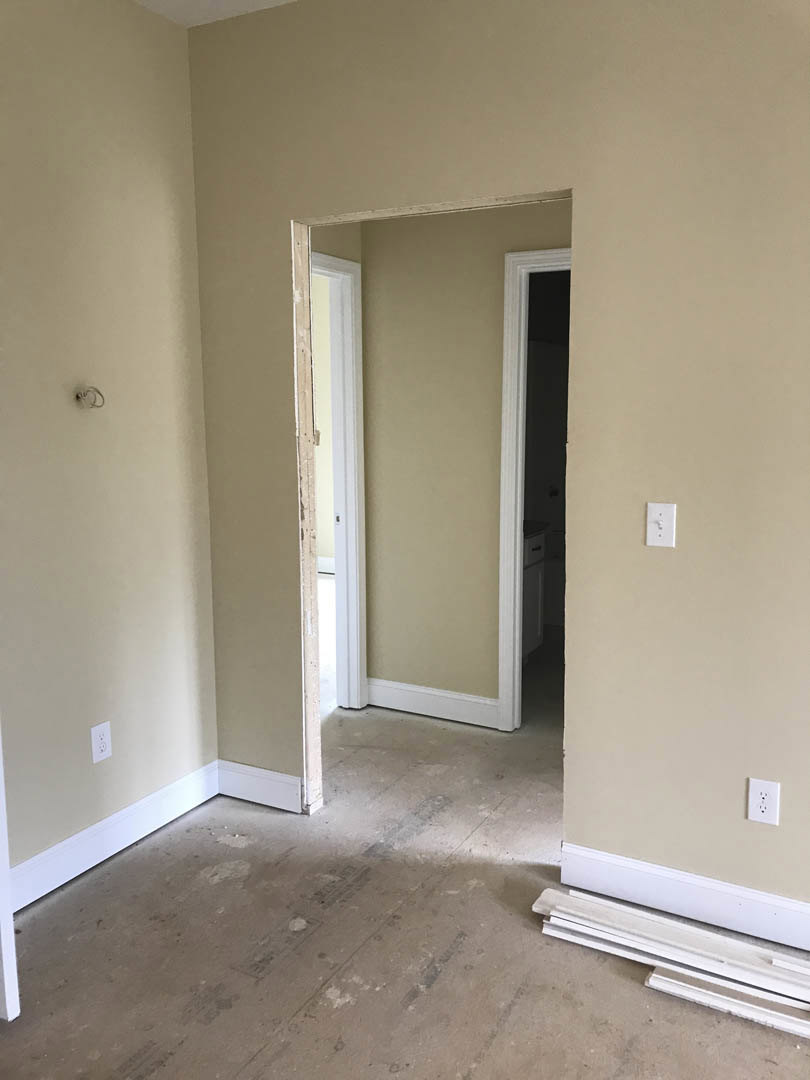 White paneled door with brown hinges open into a room with white painted walls and floor, simple molding, and minimal decor.